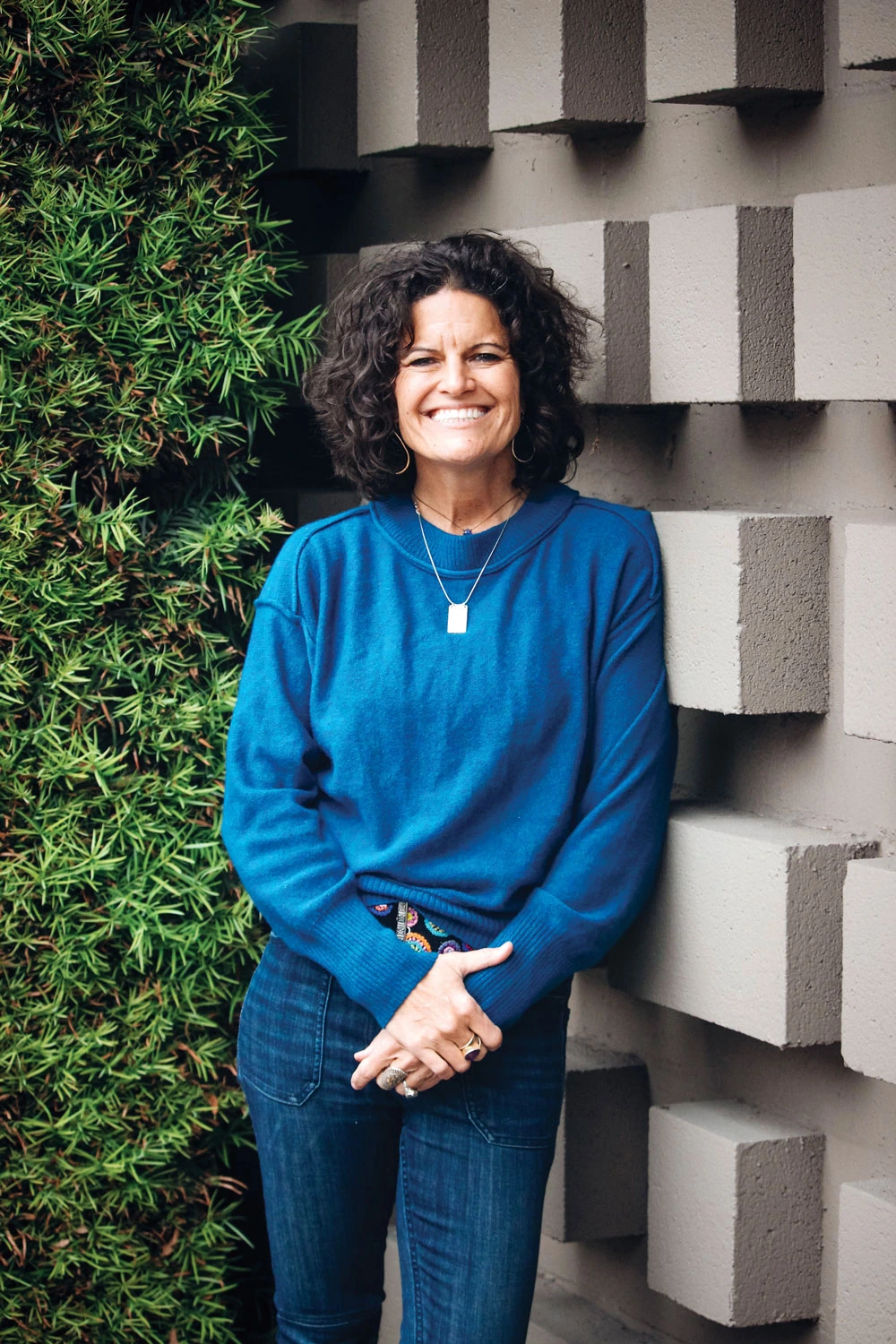 Dr. Mindy Pelz smiling in a blue sweater standing outdoors beside a textured wall and greenery.