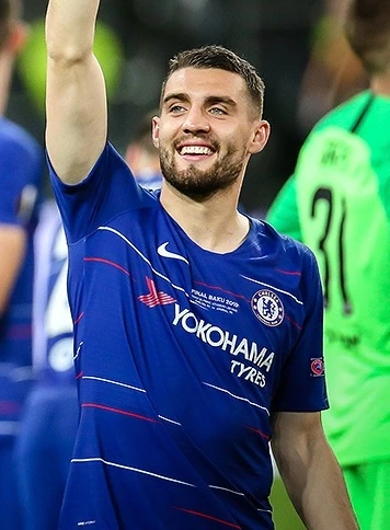 Mateo Kovačić in a Chelsea match, smiling and raising his arm in a blue jersey.