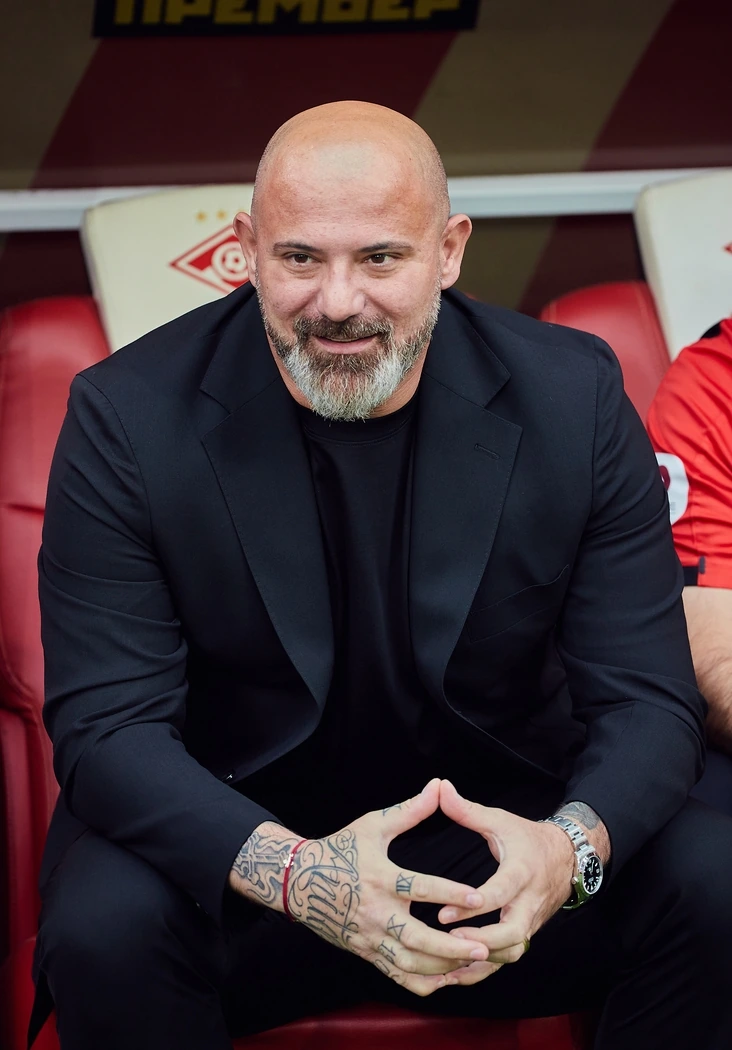 Dejan Stanković seated on the bench in a black suit, smiling during a football match