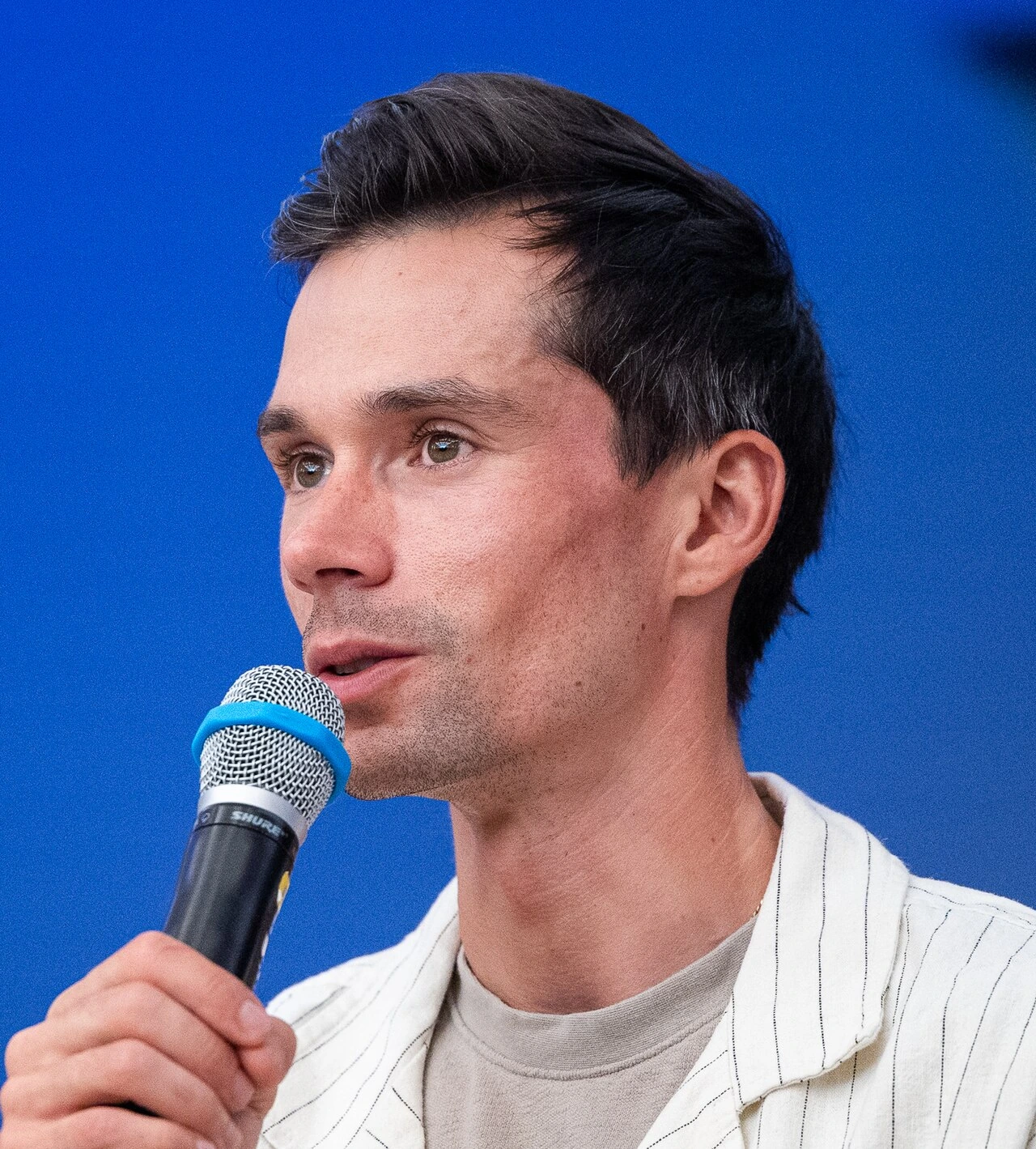 Primož Roglič speaking into a microphone at an event, close-up portrait against a blue background.