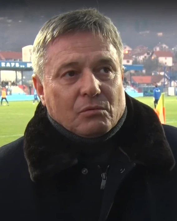 Dragan Stojković Piksi standing on a football pitch during an interview, wearing a dark coat with stadium lights and a b