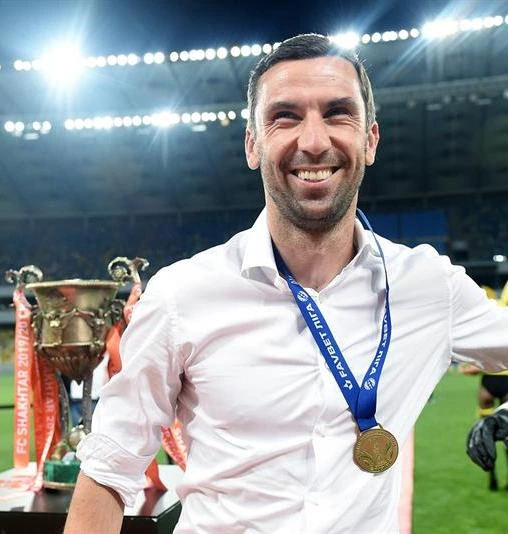 Darijo Srna smiling on a football pitch after a match, wearing a medal and white shirt with stadium lights in the backgr