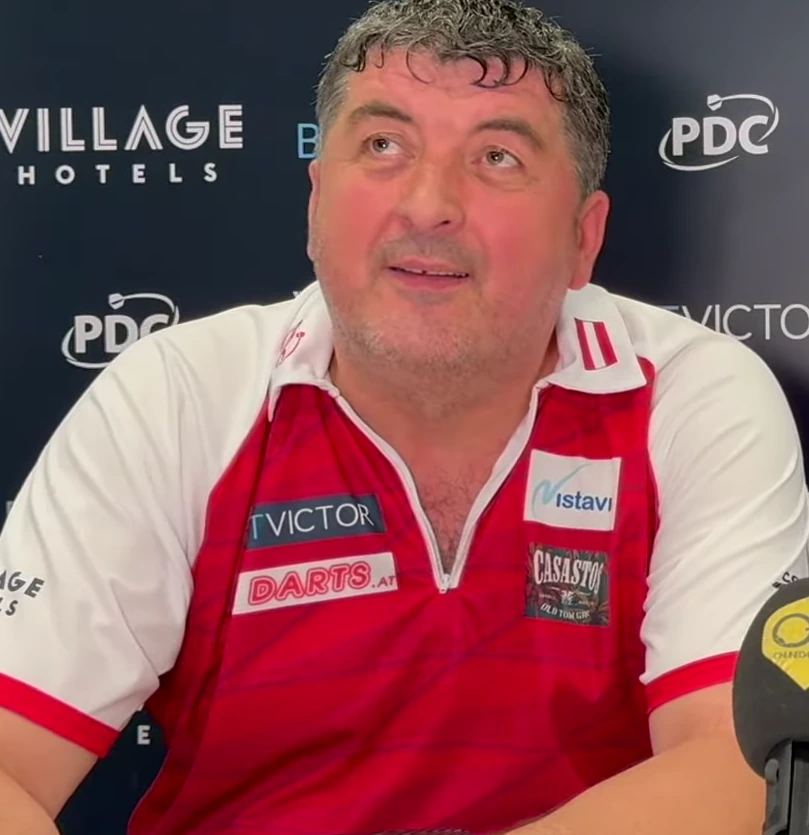 Mensur Suljović in a close-up interview-style photo wearing a red-and-white darts shirt.