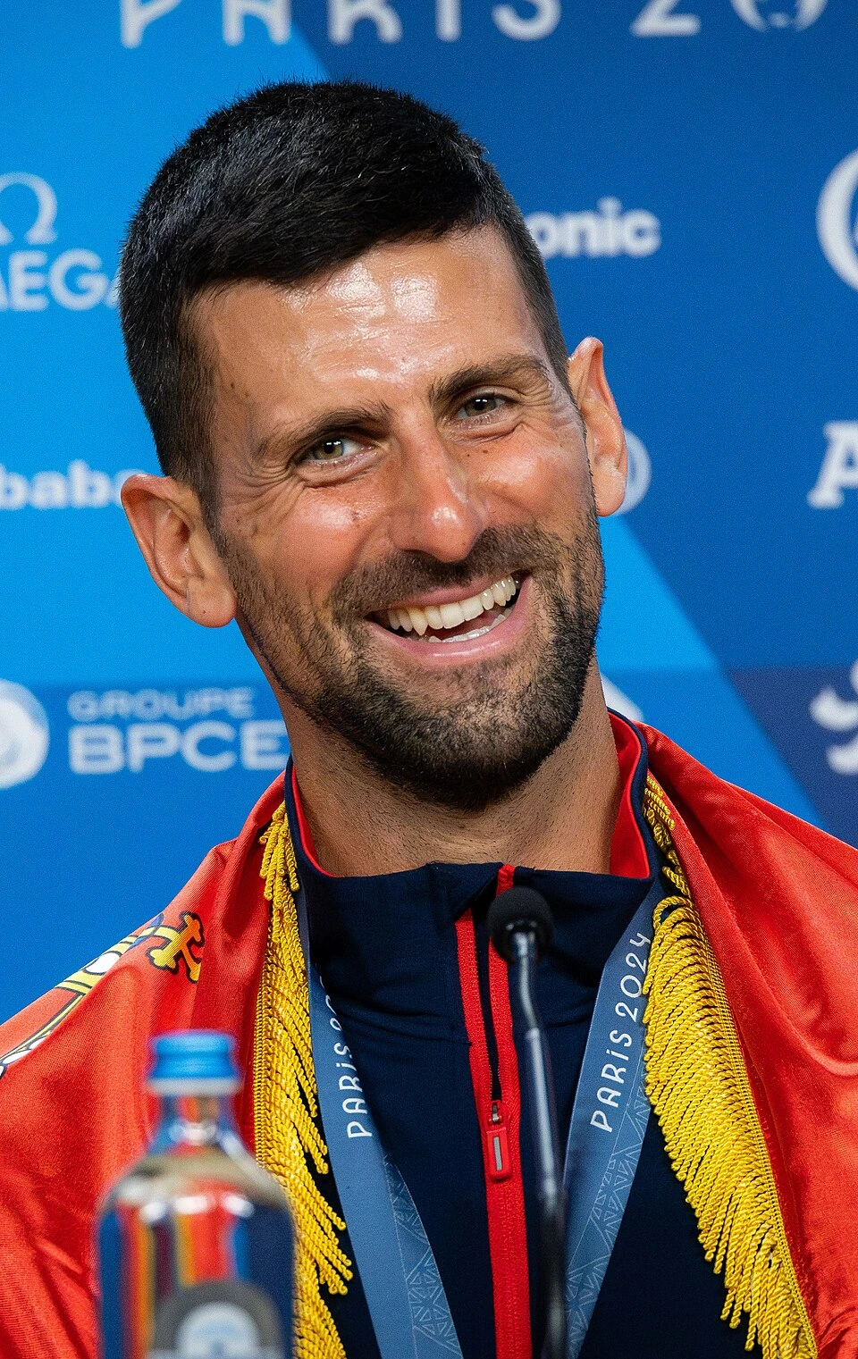 Novak Djokovic smiling at a press conference with a medal and Serbian flag draped over his shoulders