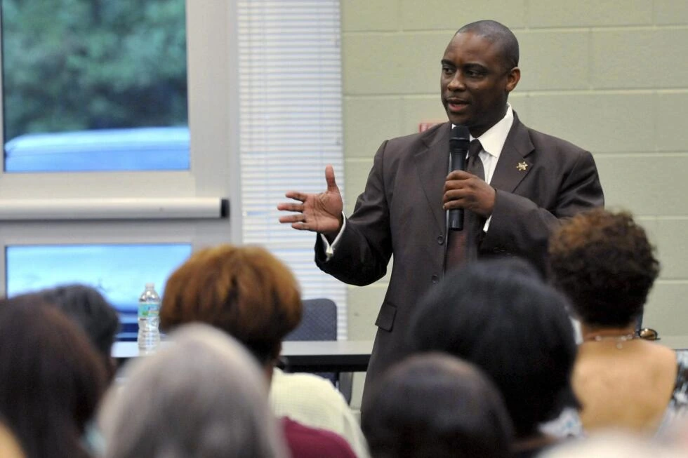 Victor Hill speaking at an event in a suit, holding a microphone before an audience