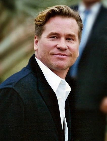 Val Kilmer at Cannes, wearing a dark suit and white shirt, turning toward the camera.