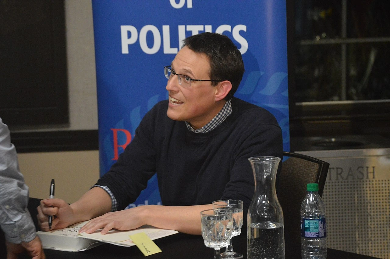 Steve Kornacki speaking at an event while seated at a table with a blue politics backdrop behind him