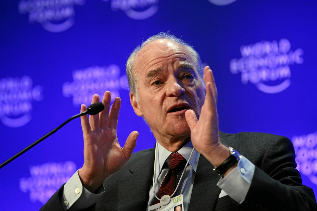 Henry Kravis speaking at an event, gesturing beside a microphone against a blue background.
