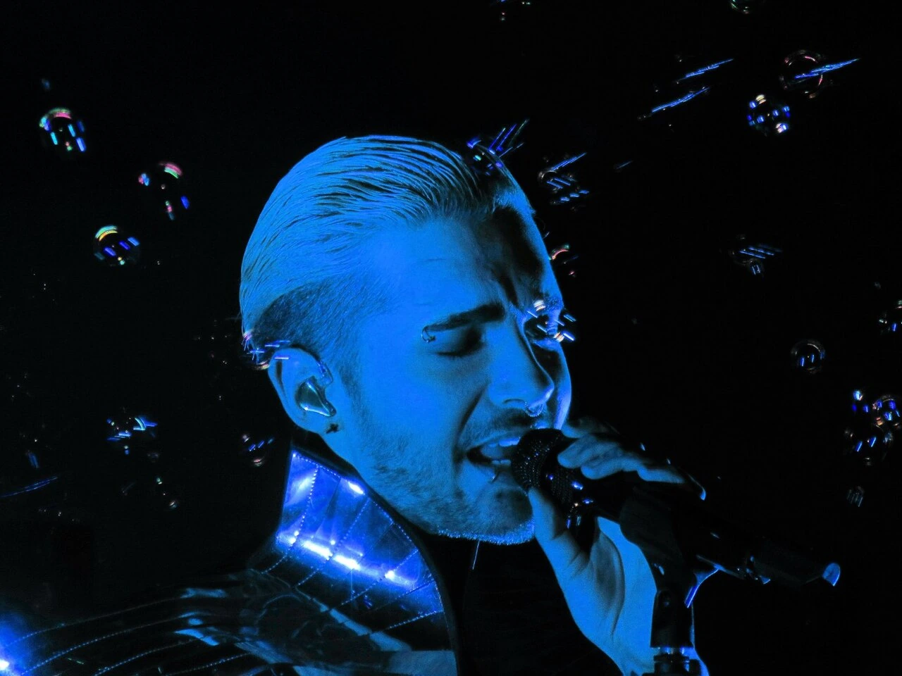 Bill Kaulitz singing into a microphone under blue stage lighting.