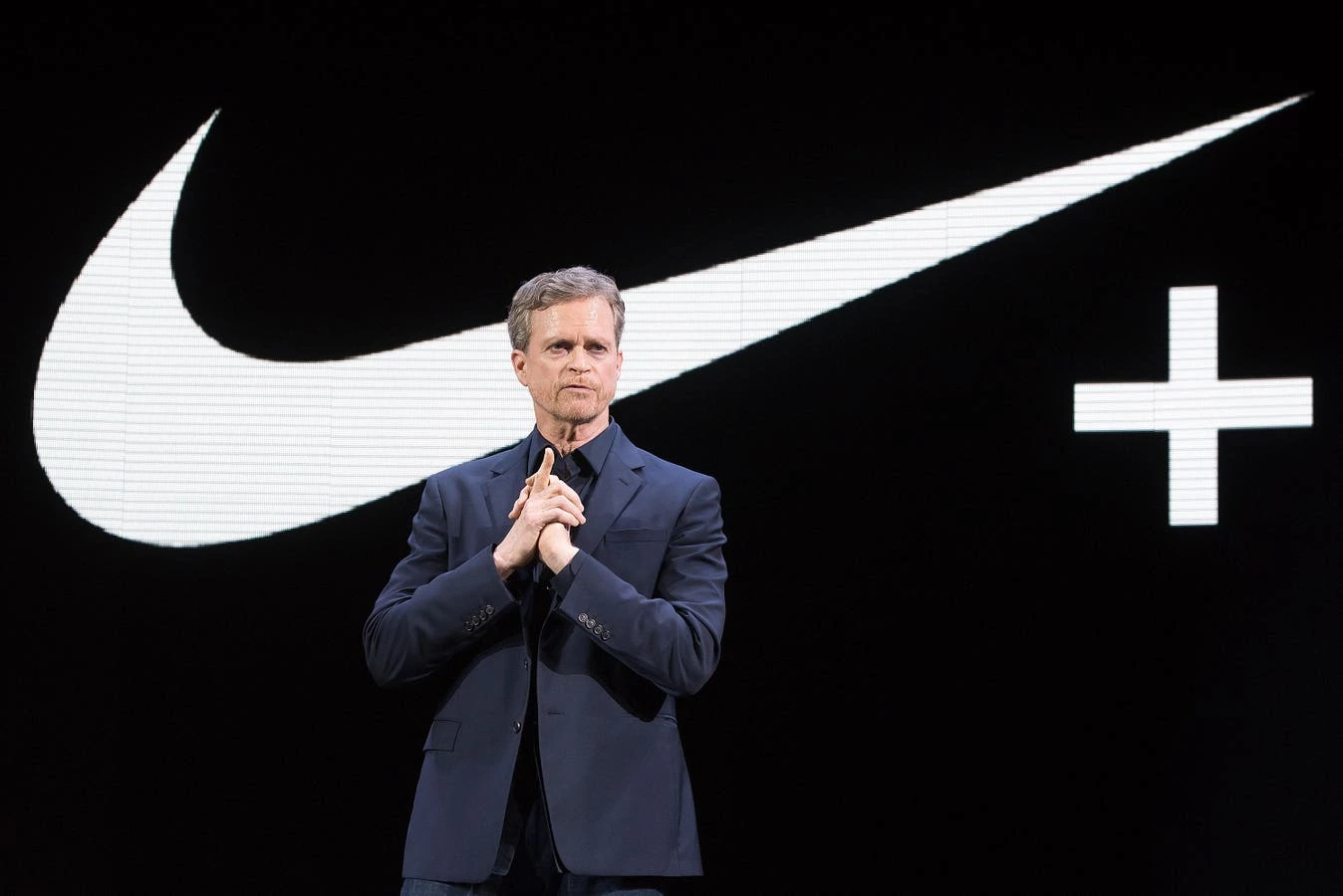 Mark Parker standing on stage in front of a large Nike logo, wearing a dark suit and clasping his hands.