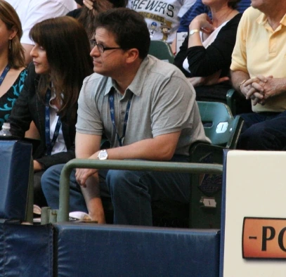 Mark Attanasio seated at a Milwaukee Brewers game, wearing glasses and a gray polo shirt.