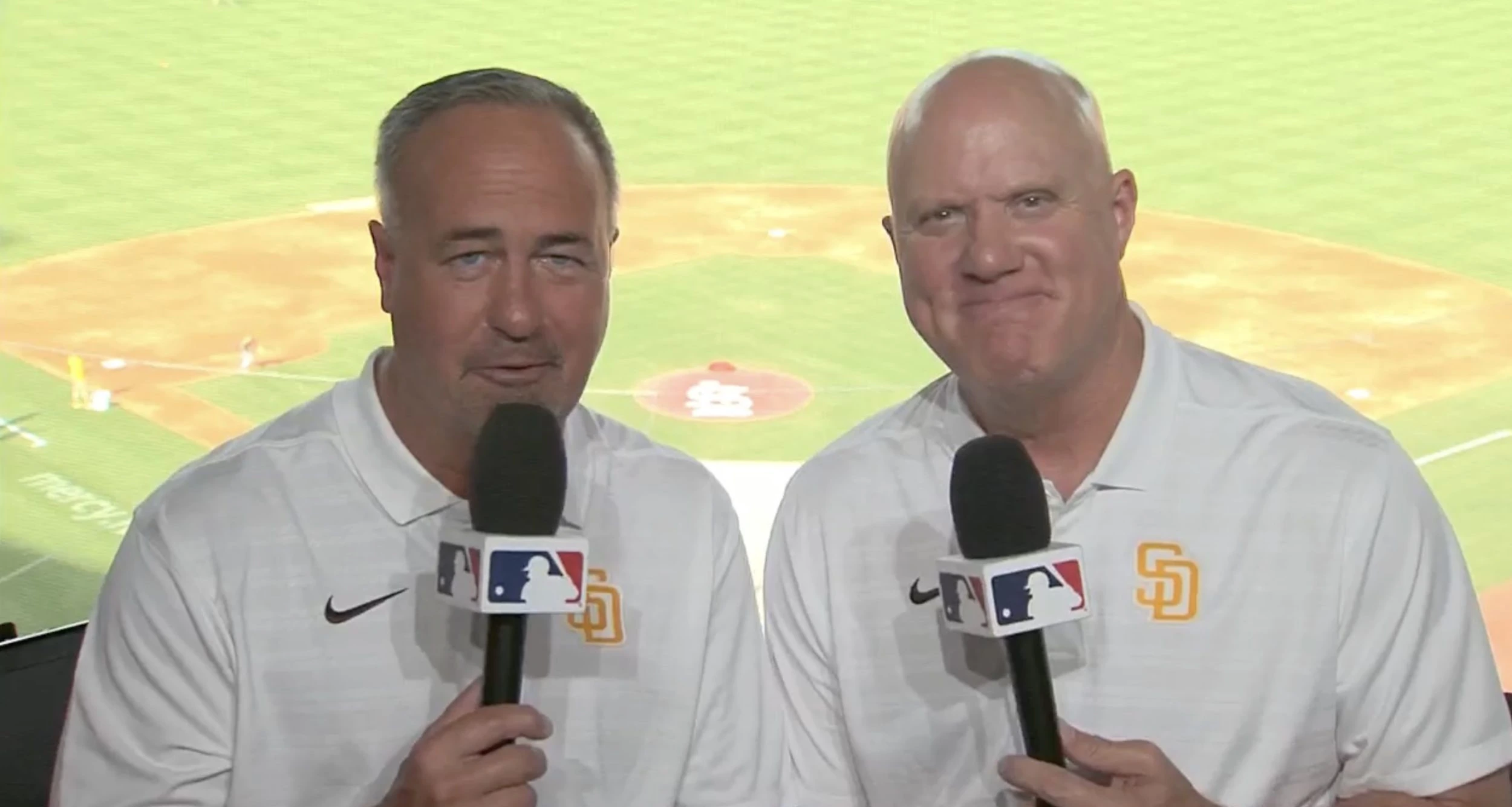 Mark Grant speaking as a Padres broadcaster on camera with a microphone in front of a baseball field backdrop.