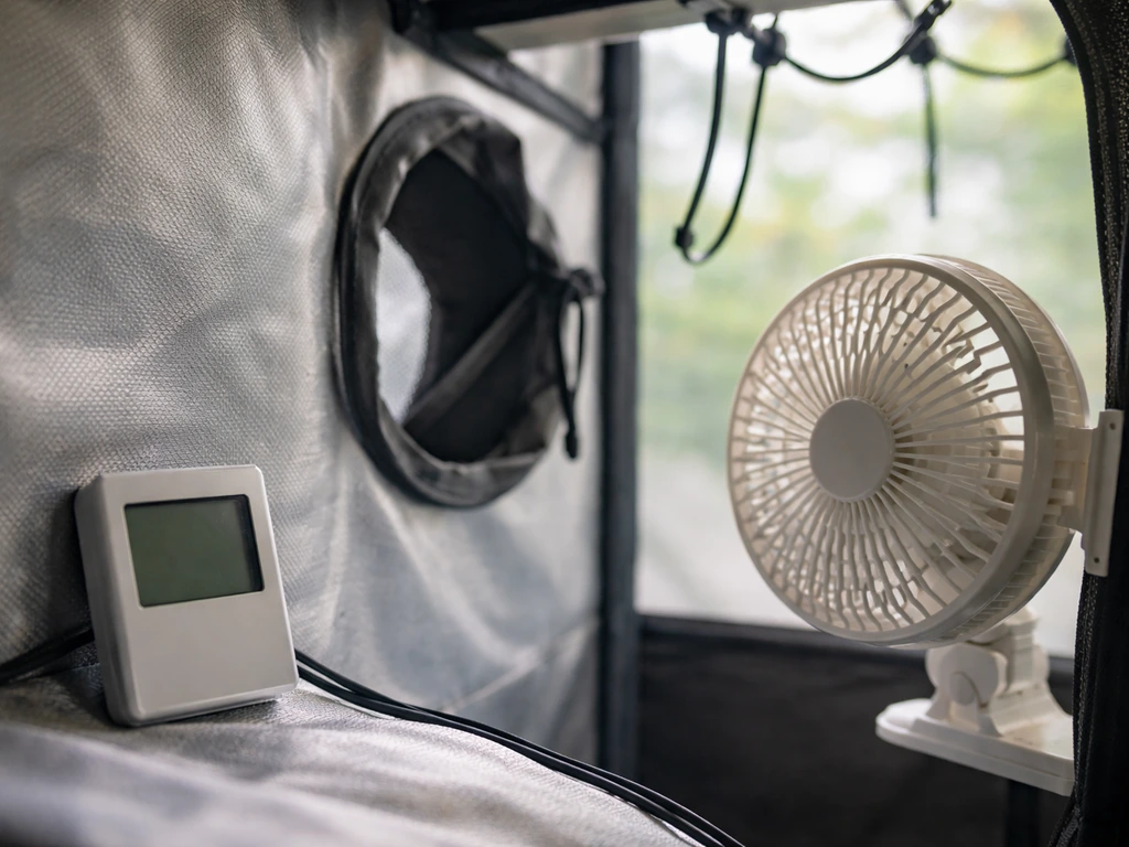 Close-up thermometer probe inside a grow tent next to a vent and small fan for temperature safety checks.