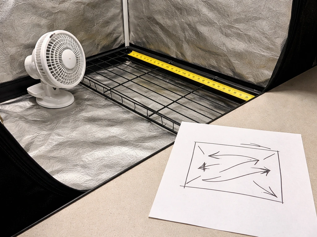 Close-up of an airflow calculation setup: tape measure, paper sketch, and a small fan beside a grow-tent interior.