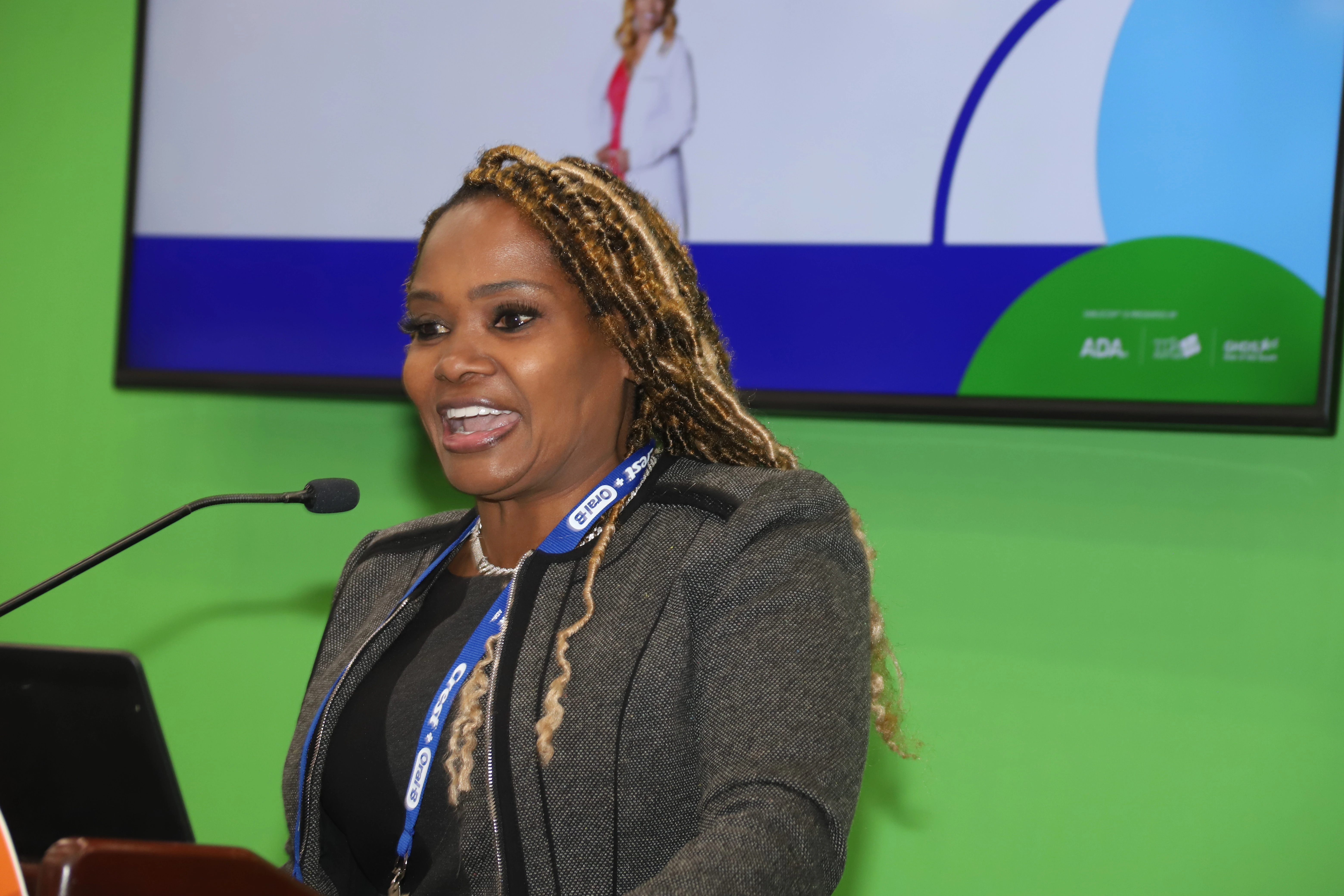 Heavenly Kimes speaking at a podium during a dental event, with a microphone in the foreground and a colorful presentati