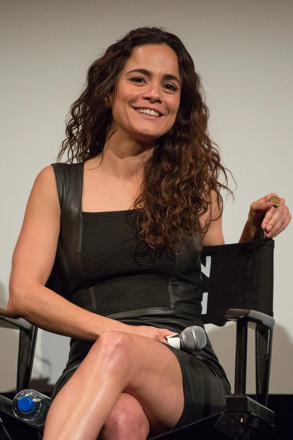 Alice Braga smiling while seated at a Queen of the South event, holding a microphone.