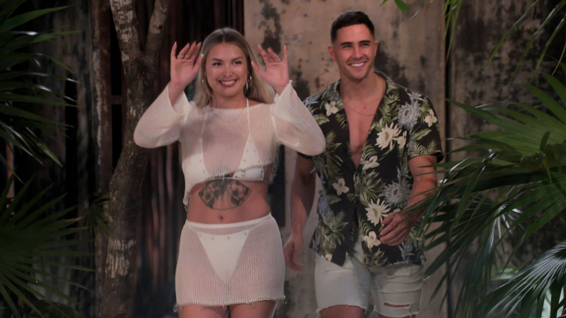A smiling woman and man from Love Is Blind walk outdoors through tropical foliage.
