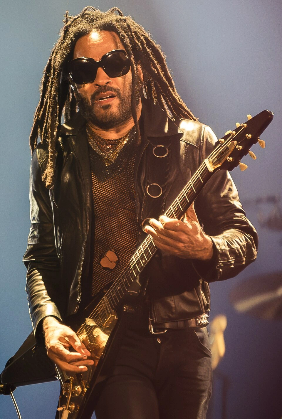 Lenny Kravitz performing on stage with a guitar, wearing sunglasses and a leather jacket.
