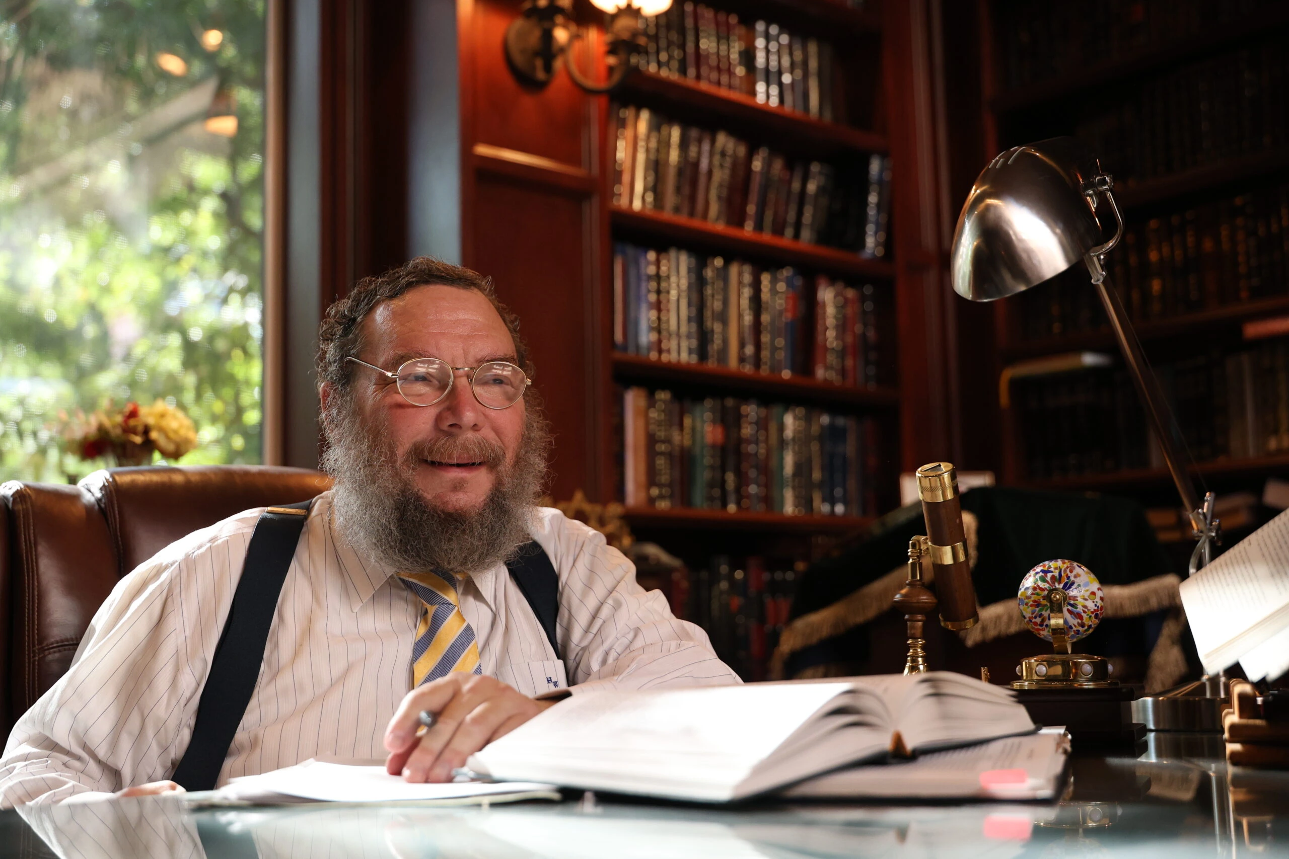 Rabbi Moshe Weiss seated at a desk in a study lined with books, looking off to the side.