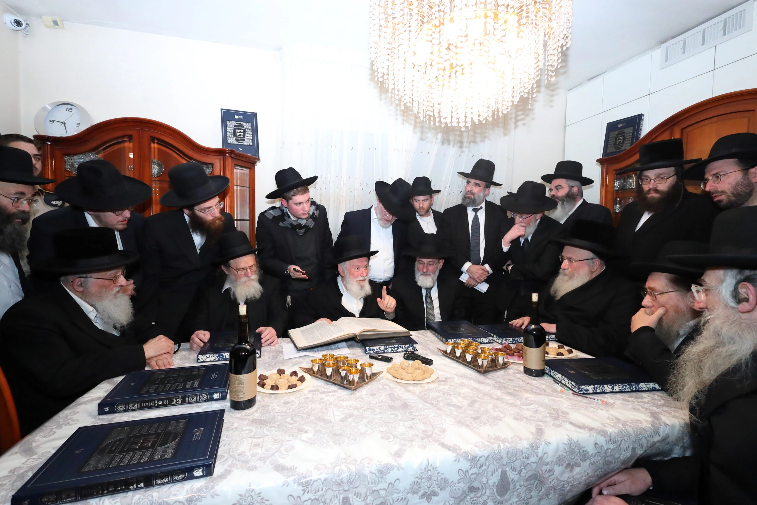 Rabbi Dovid Hofstedter seated at a table during a Torah gathering, wearing a black hat and suit, speaking among other me