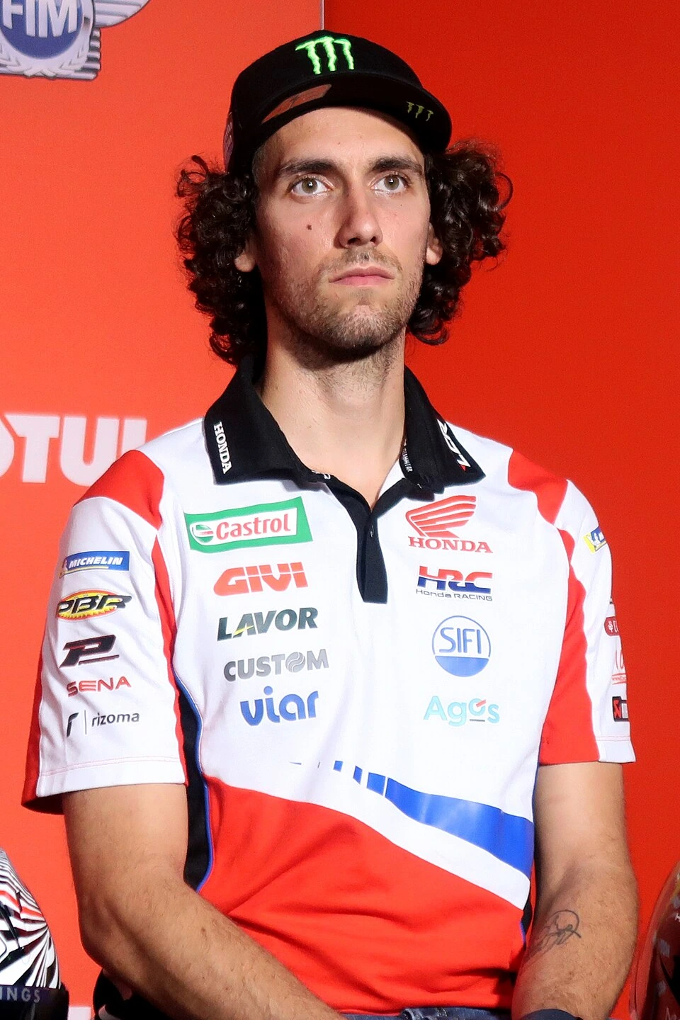 Álex Rins in a Honda racing shirt and black cap, posing against a red backdrop