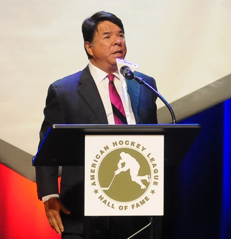 Ray Halbritter speaking at a podium during an event, wearing a dark suit and pink tie.