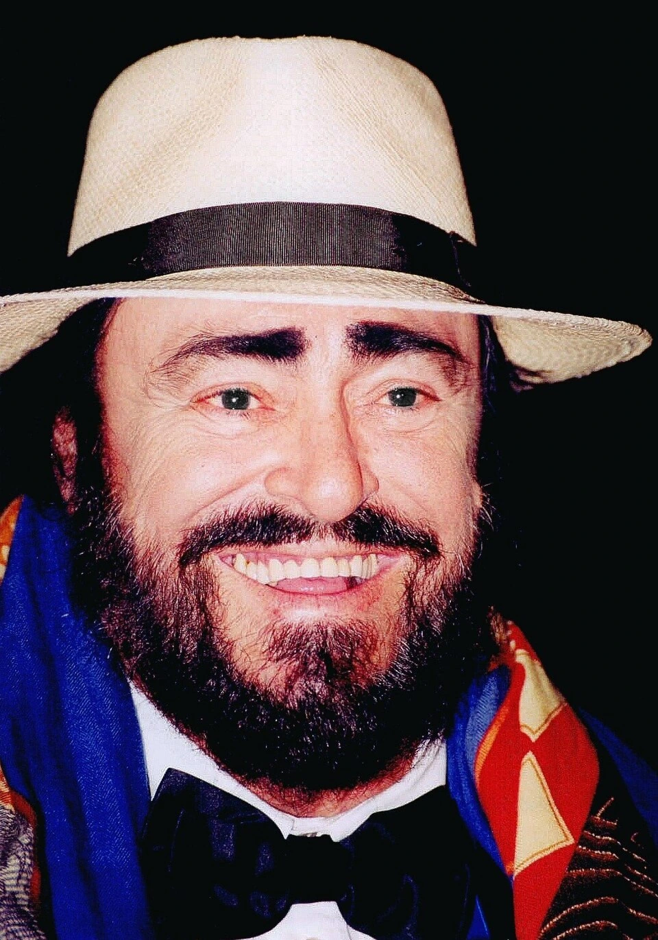 Luciano Pavarotti smiling in a close-up portrait wearing a light-colored hat and colorful scarf