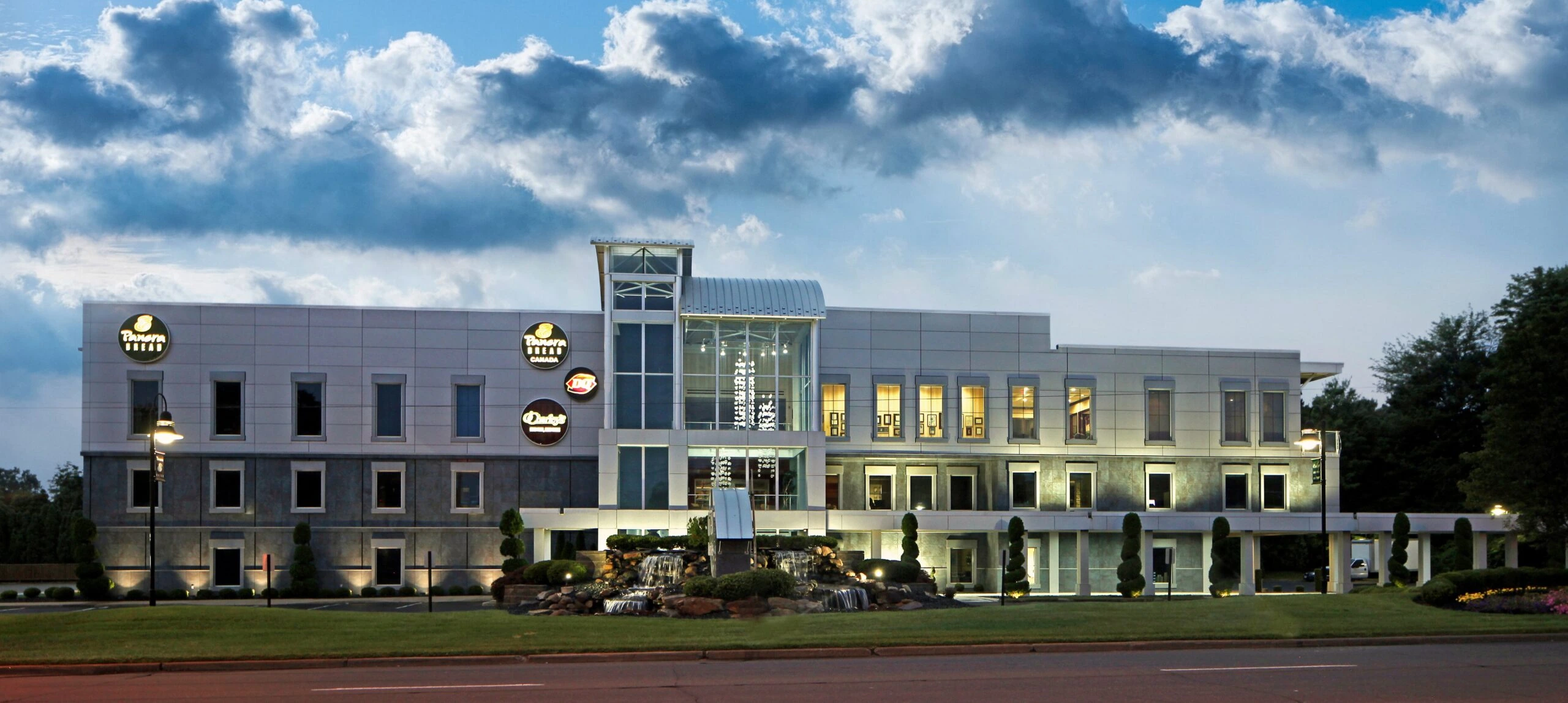 A modern Covelli Enterprises office building at dusk with illuminated windows, landscaping, and a fountain in front.