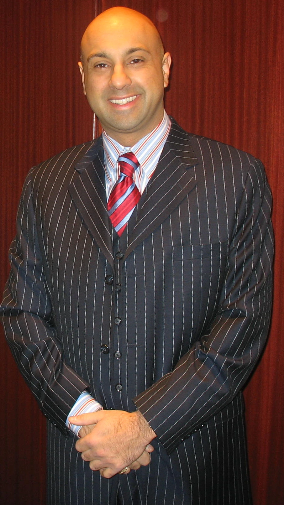 Ali Velshi in a pinstripe suit and tie, standing indoors and smiling at the camera.