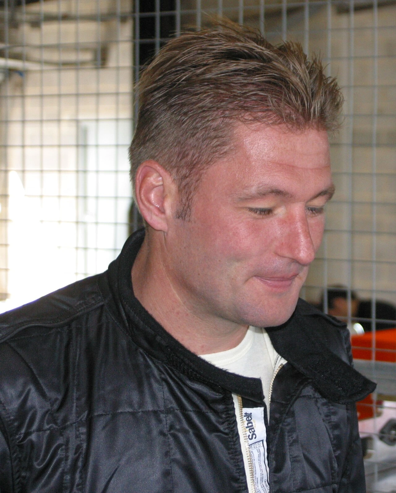 Jos Verstappen in a racing jacket, looking downward in a candid close-up photo.