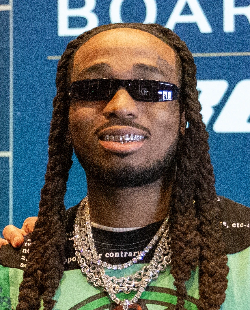 Quavo wearing sunglasses and layered chains at an event backdrop.