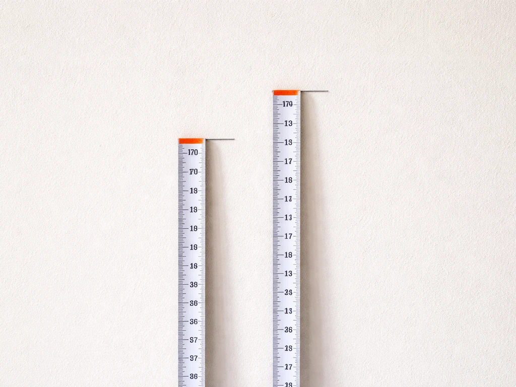 Two measuring tapes against a plain wall showing a small 3–6 cm height difference.
