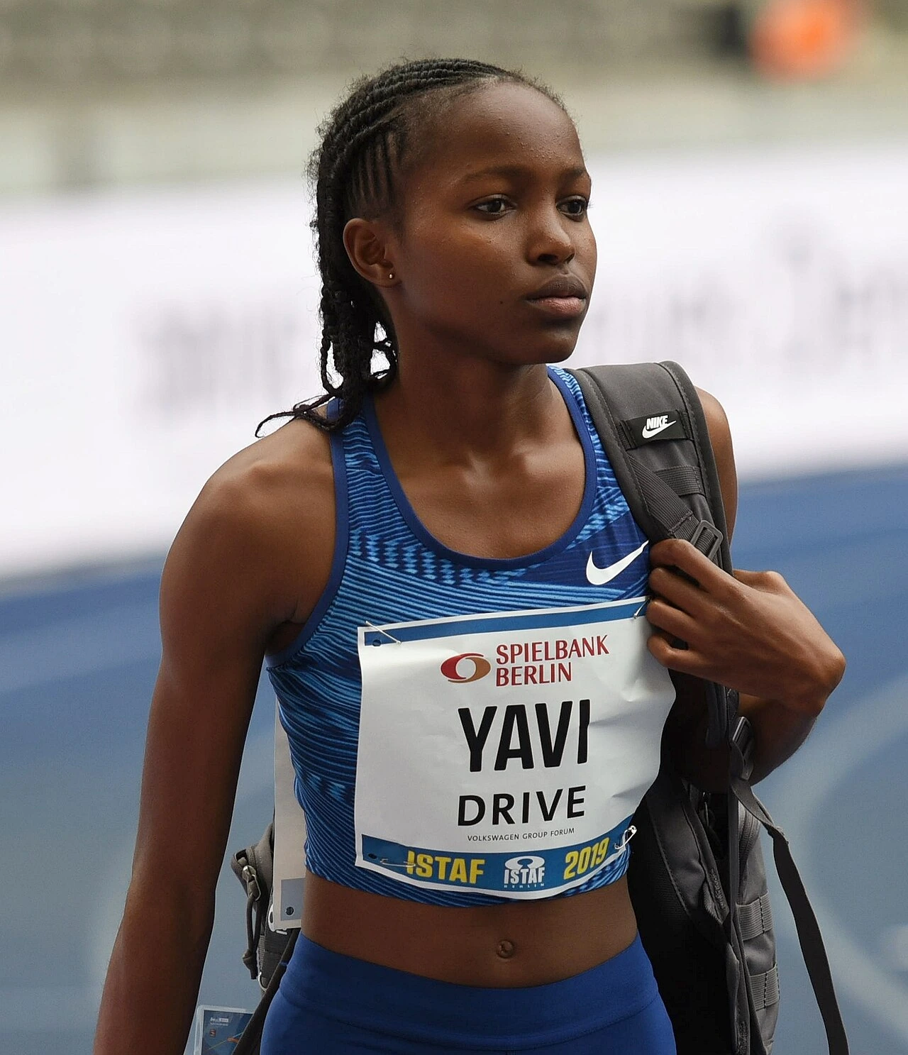 Winfred Mutile Yavi standing at a track meet wearing a blue racing kit and bib, carrying a backpack