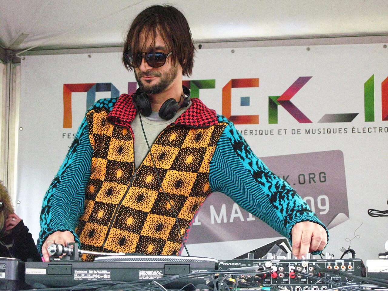 Ricardo Villalobos DJing on stage at a festival, wearing sunglasses and headphones around his neck.
