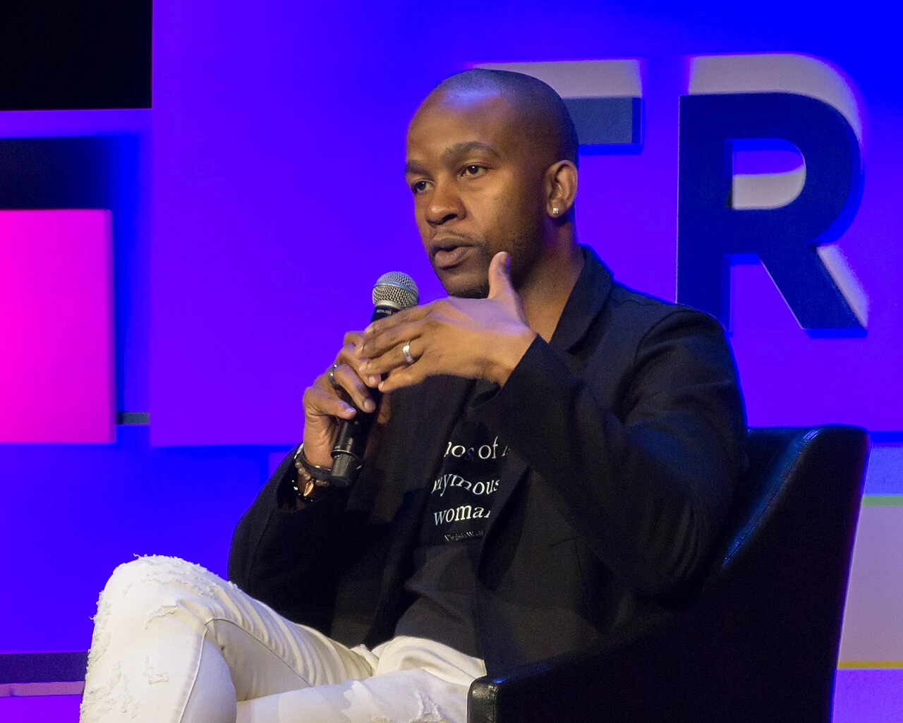 Wade Davis speaking on stage at a conference, holding a microphone and seated in a chair against a lit backdrop.