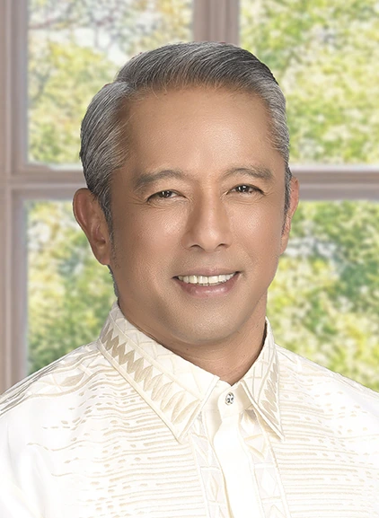 Portrait photo of Jonvic Remulla smiling in a formal shirt, posed indoors near a window with a blurred outdoor backgroun