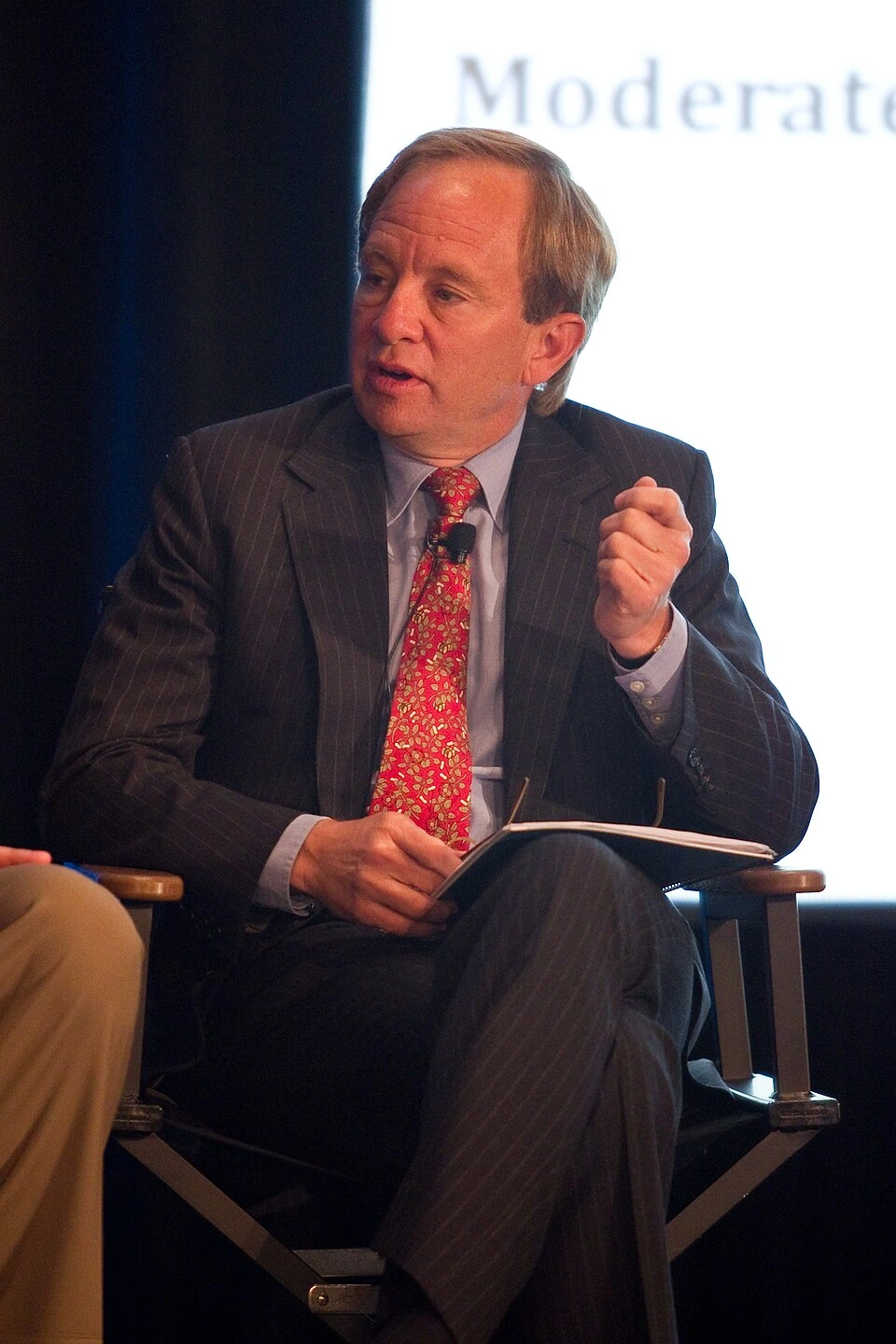 Steven Rattner speaking at an event in a suit and red tie, seated on stage.