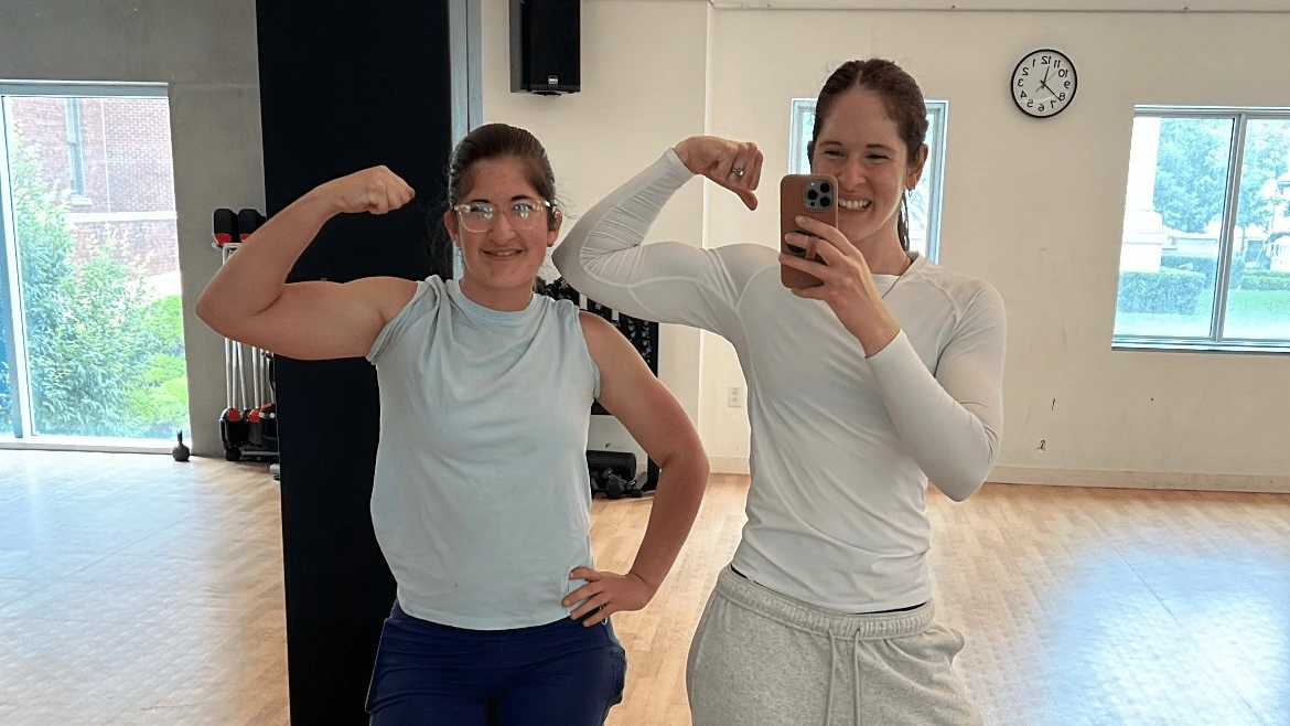 Two women posing in a fitness studio, flexing their arms in front of a mirror.