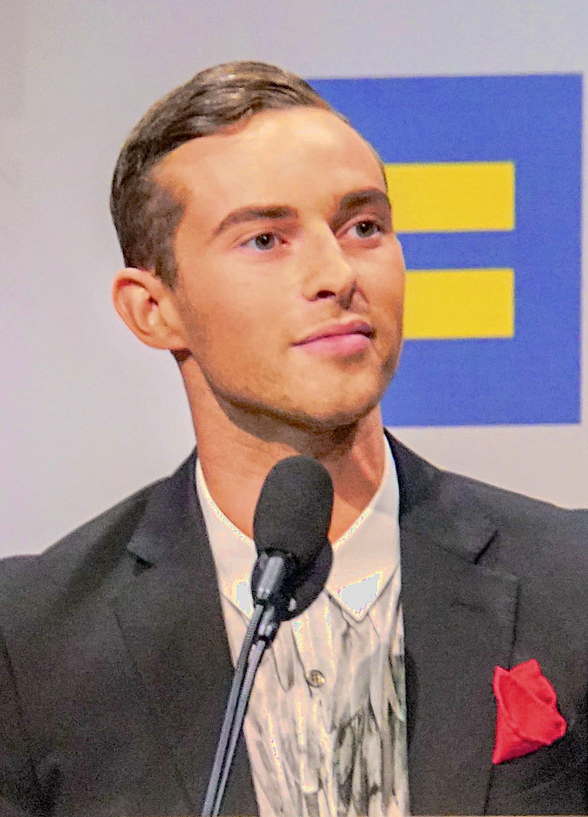 Adam Rippon speaking at an event, wearing a suit with an Equality logo in the background