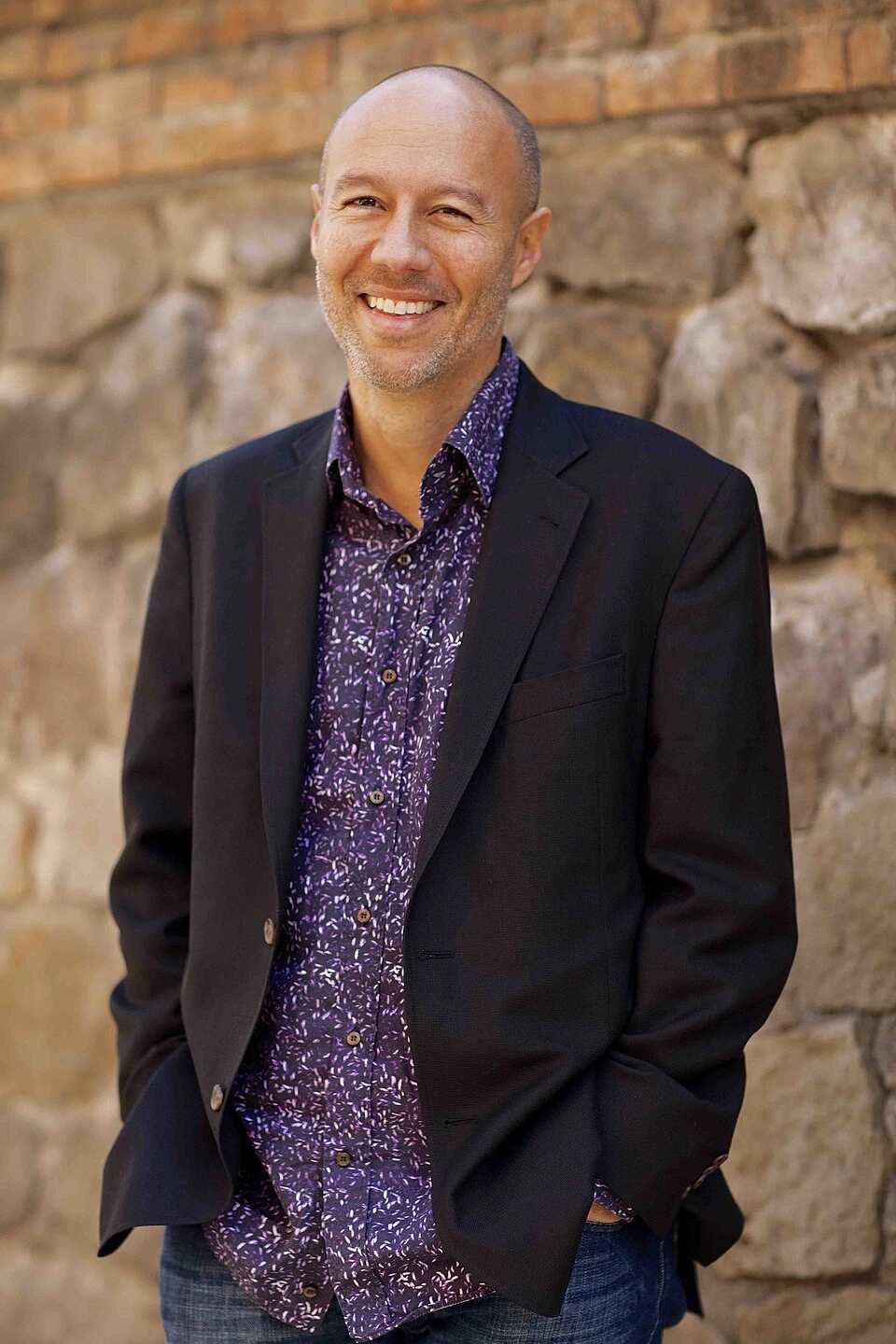 David Risher smiling in a portrait-style photo against a stone wall background