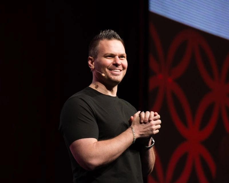 John Ruhlin speaking on stage with a headset microphone, smiling in a black shirt against a dark event backdrop.