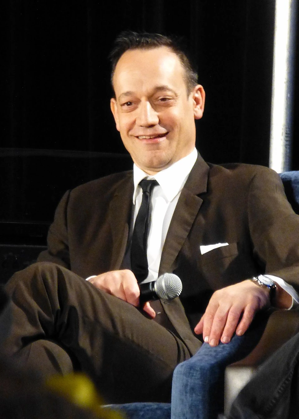 Ted Raimi seated at an event, holding a microphone and wearing a suit.