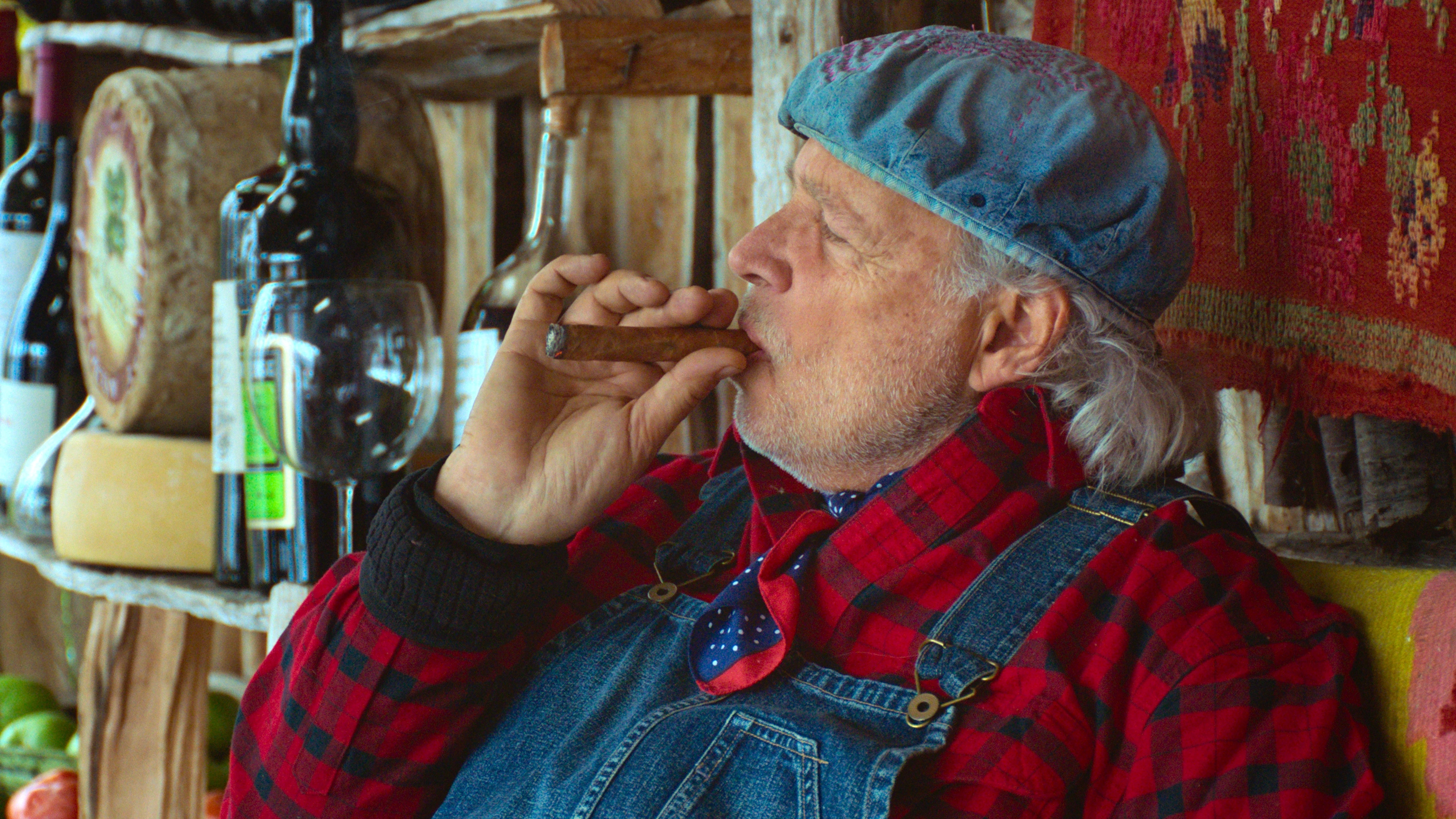 Francis Mallmann smoking a cigar in a rustic setting, wearing a cap and red plaid shirt under denim overalls.