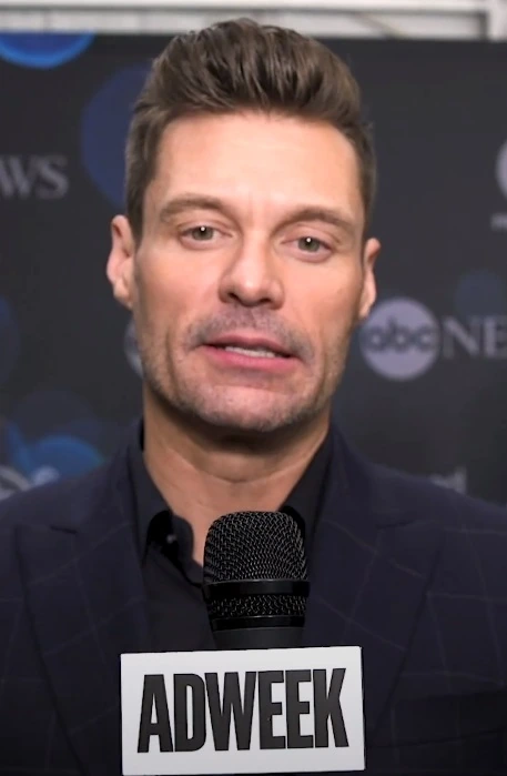 Ryan Seacrest speaking at an event with an ADWEEK microphone in front of a branded backdrop