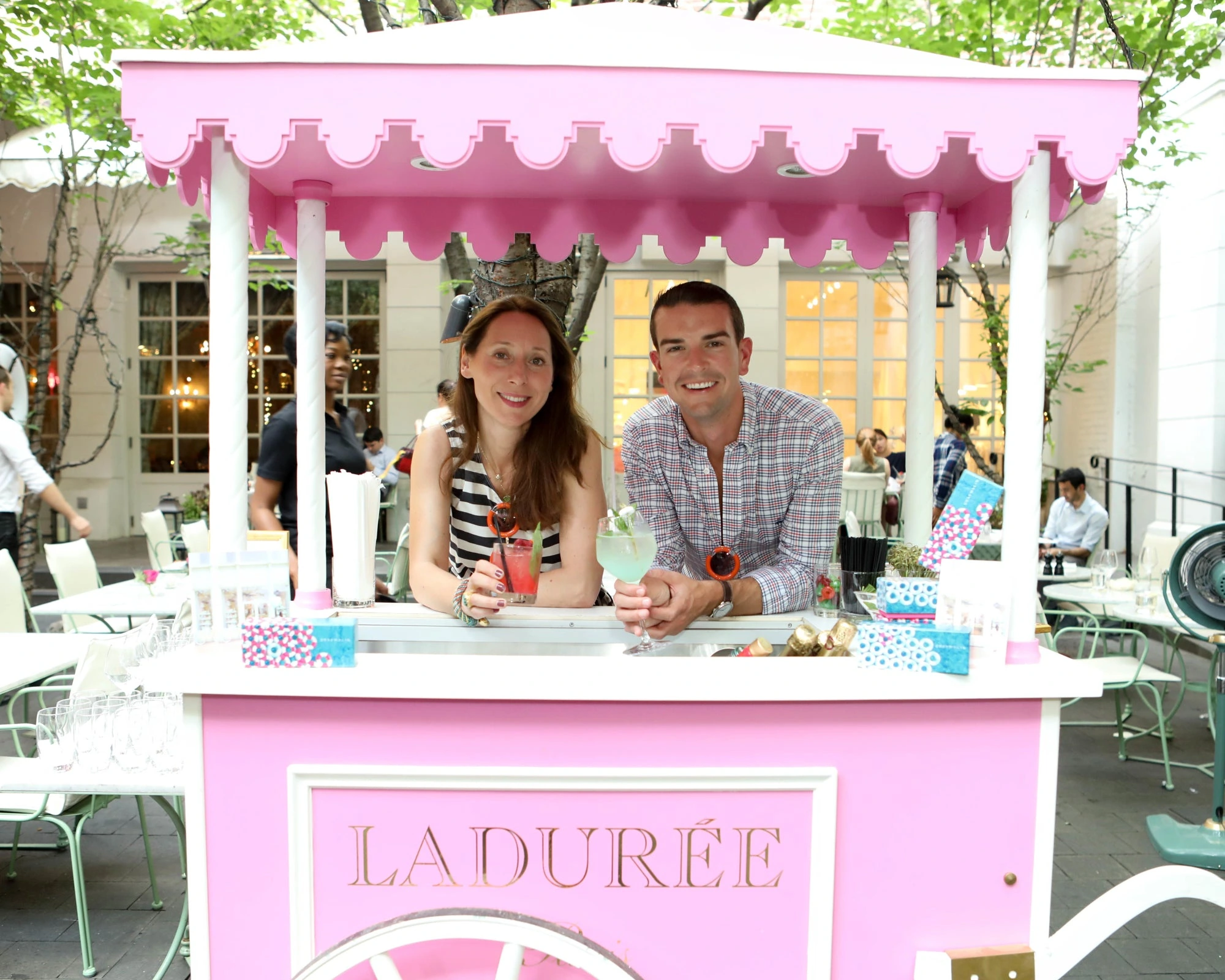 A woman and man smiling behind a pink La Durée stand at an outdoor café.