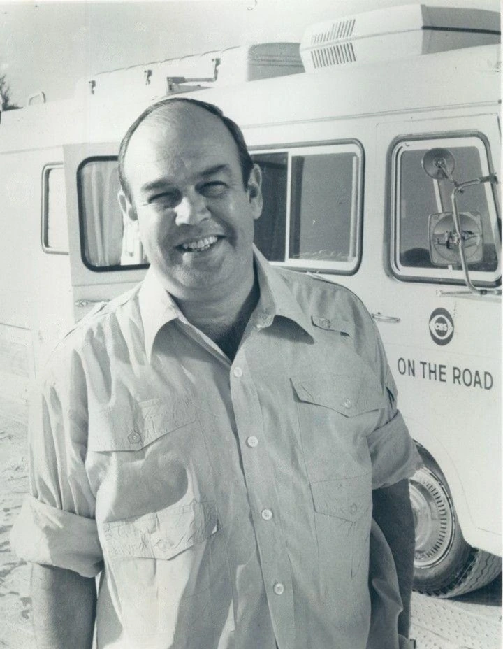 Black-and-white publicity photo of Charles Kuralt standing beside a CBS vehicle marked “On the Road”