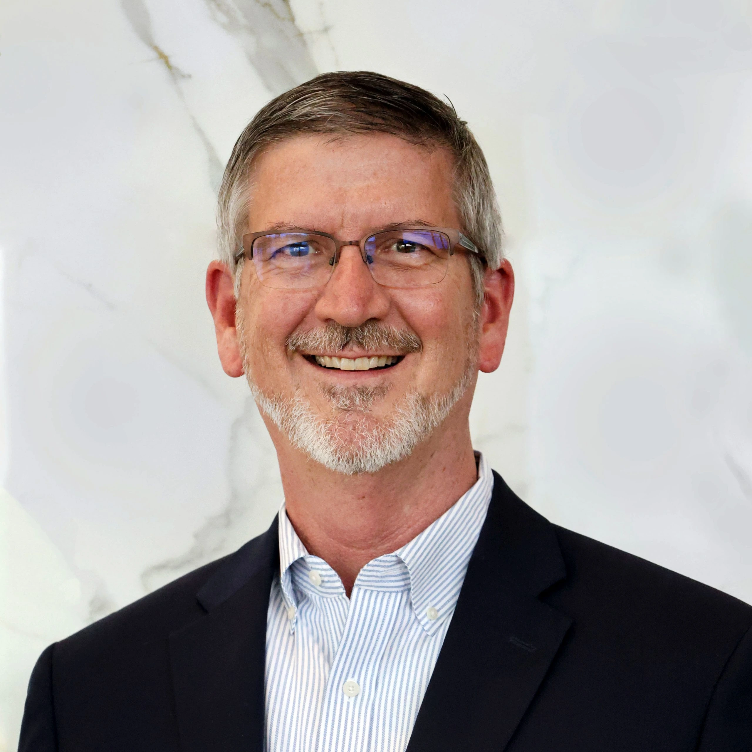 Portrait of Charles Peek, CEO of Custard Insurance Adjusters and Riverwood, smiling in a suit and glasses against a ligh
