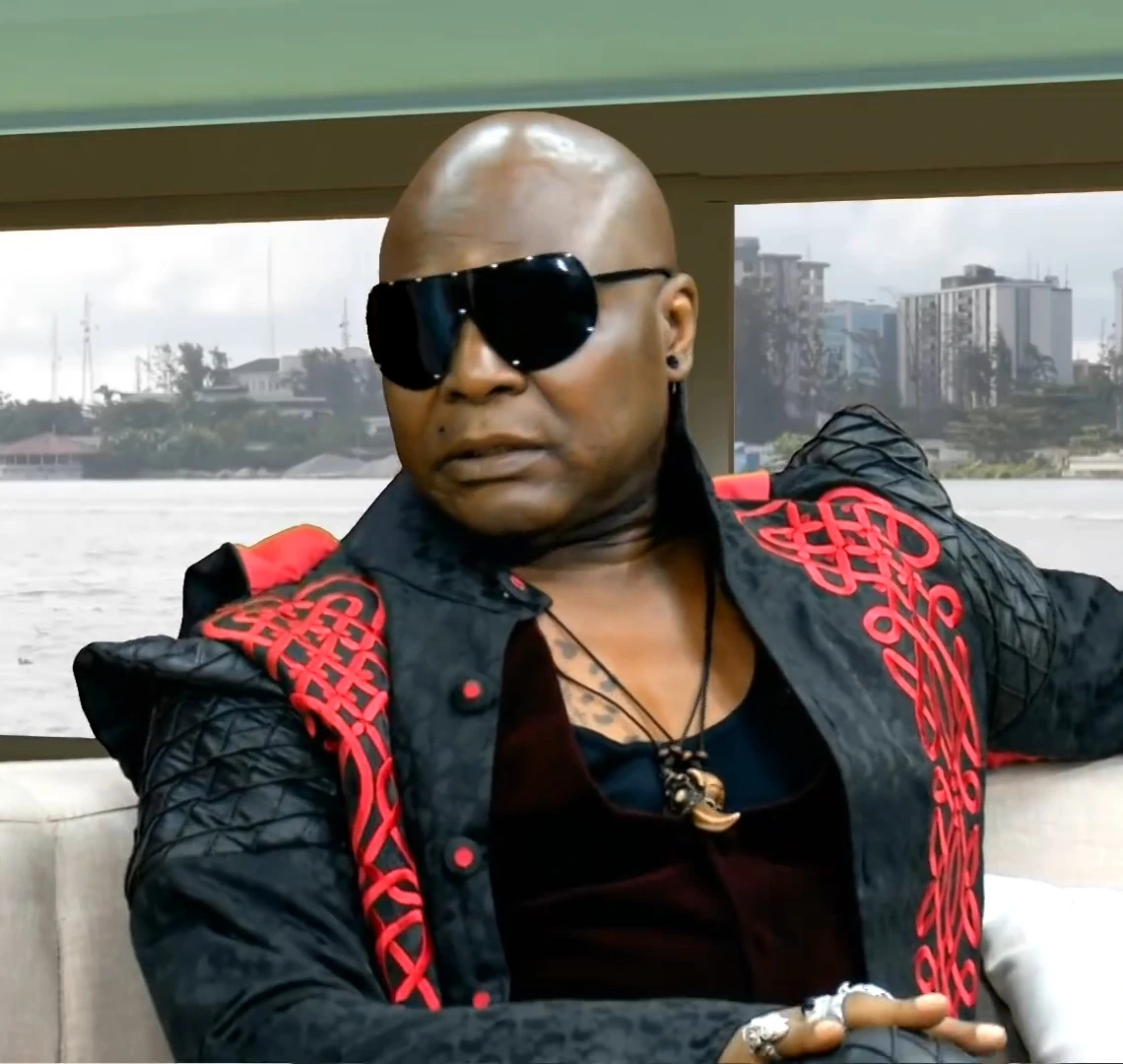 Charles Chukwuemeka Oputa (Charly Boy) seated indoors wearing sunglasses and a black-and-red outfit, with a waterfront c