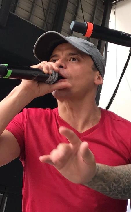 Vico C performing into a microphone while wearing a gray cap and red shirt