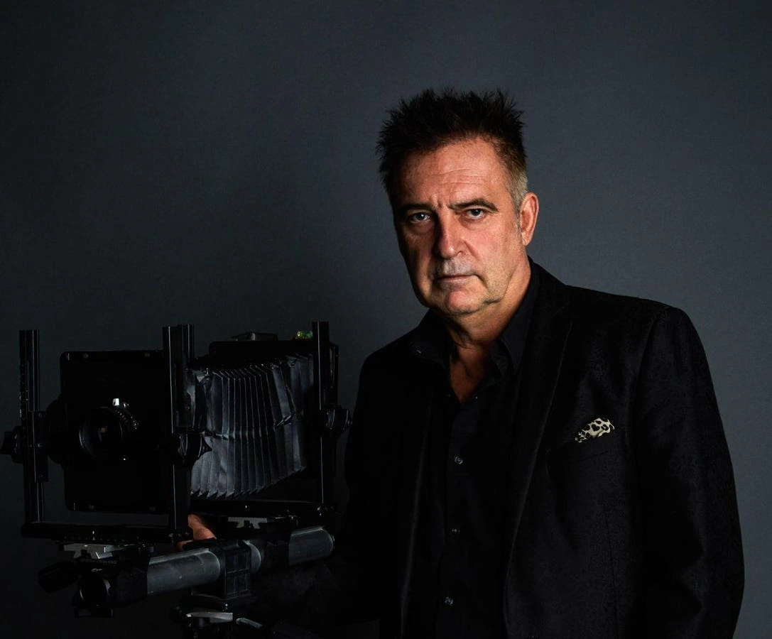 Portrait of Antoine Verglas standing beside a large-format camera against a dark studio background